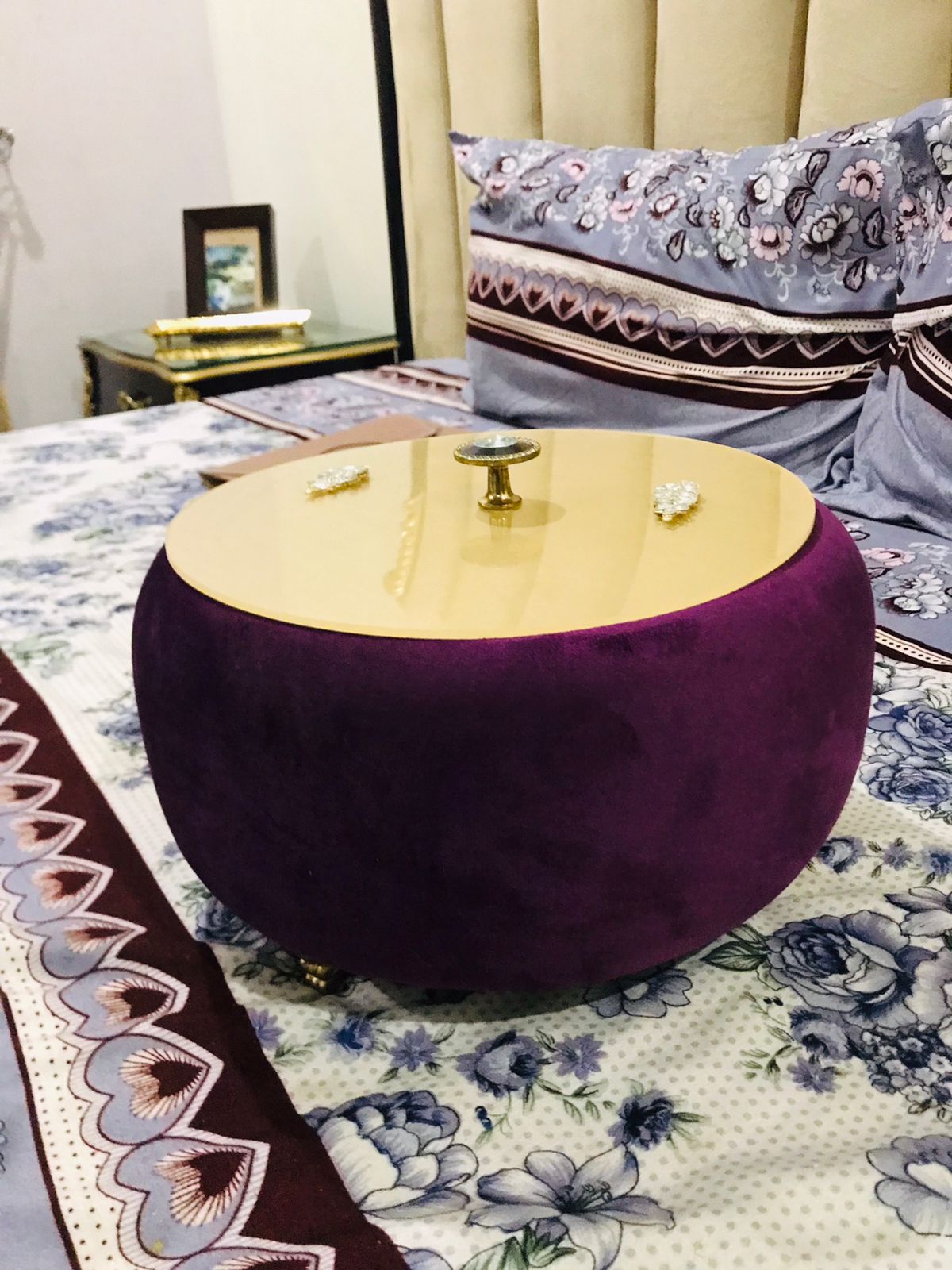Round gold Turkish Hotpot on a purple cushion with a floral bedspread and framed picture in the background